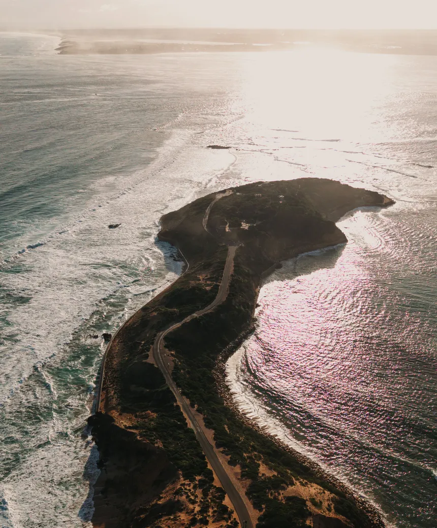 An aerial view of a narrow coastal spit at sunset, with surf on the ocean side and calm water on the other