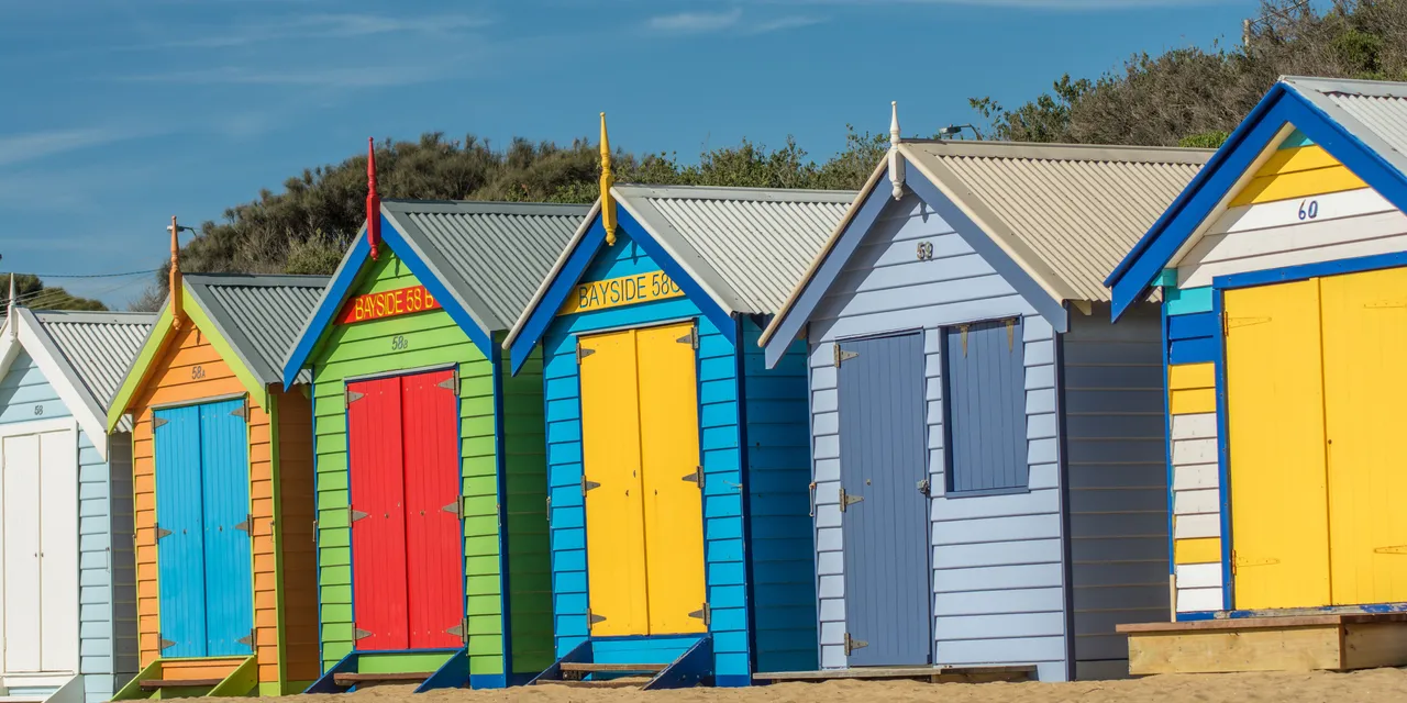 Brightly painted bathing boxes standing in a row on the sand, with scrub behind them
