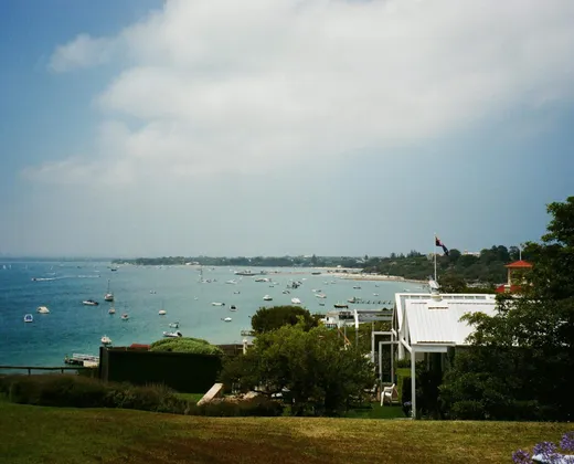 A grassy rise above a bay, with moored boats offshore and a white weatherboard house in the foreground
