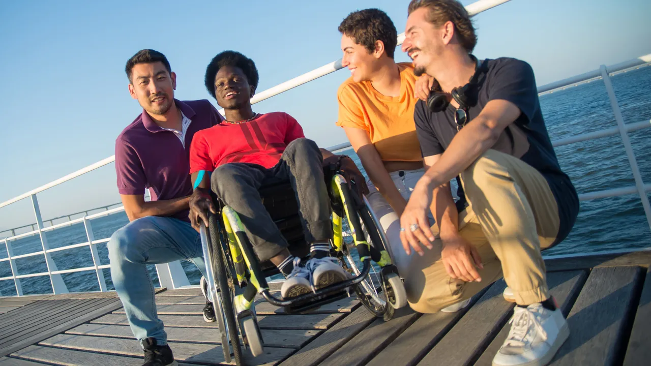 A group of friends, including a person using a wheelchair, on a boardwalk by the sea