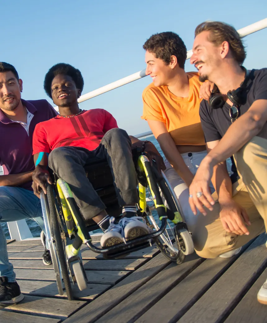 A group of friends, including a person using a wheelchair, sharing a laugh on a seaside boardwalk