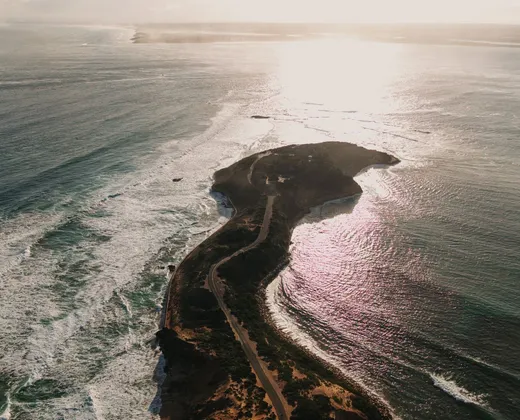 An aerial view of a narrow coastal spit at sunset, with surf on the ocean side and calm water on the other