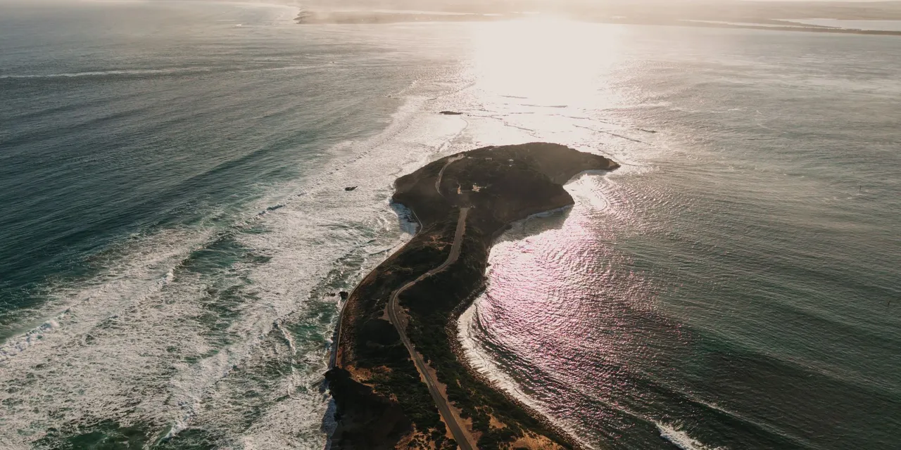 An aerial view of a narrow coastal spit at sunset, with surf on the ocean side and calm water on the other