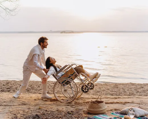 Two people on a sandy shore at sunset, one using a wheelchair, with a picnic rug and basket laid out nearby